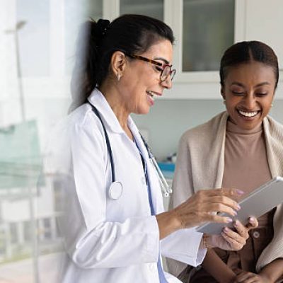 Happy senior practitioner woman and cheerful young African patient woman watching examination medical report on tablet computer, using digital technology, laughing
